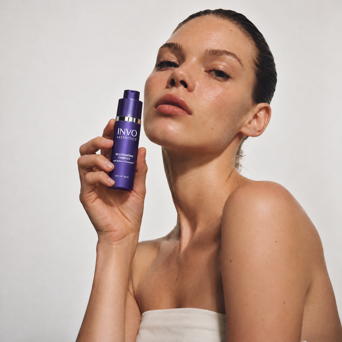 Woman holding INVO Rejuvenating Complex exosome serum beside her face in a clean studio